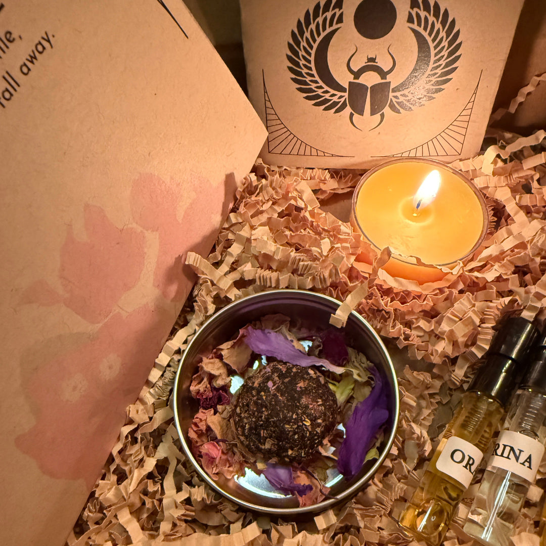 Candle, bowl with herbs and flowers, and bottles on shredded paper with a branded box in the background.
